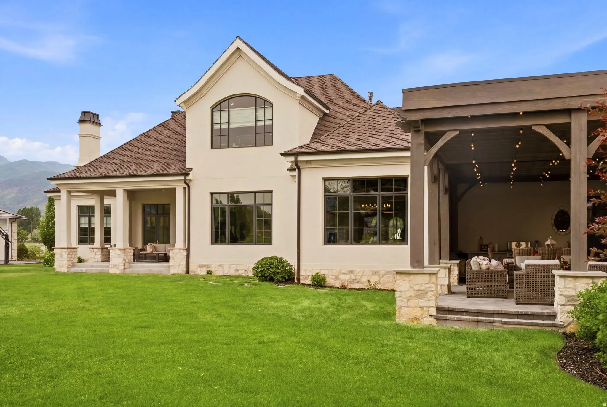 Expansive rear exterior featuring a covered patio with integrated lighting, a separate covered porch, a stucco and stone facade, dark-framed windows, and a manicured lawn