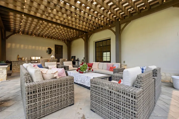 Expansive outdoor patio featuring a wood pergola with string lighting, stone-look paver flooring, a built-in outdoor kitchen with grill, and a dark-framed window