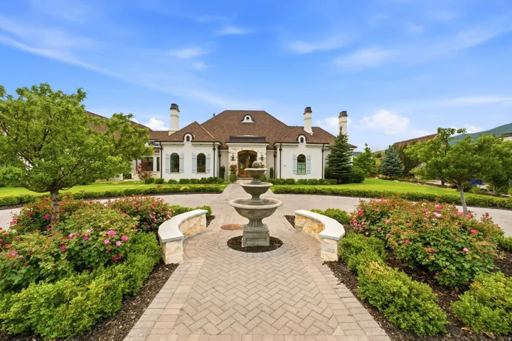 Circular paver driveway featuring a tiered stone fountain and meticulously maintained landscaping with mature trees, shrubs, and flowering bushes