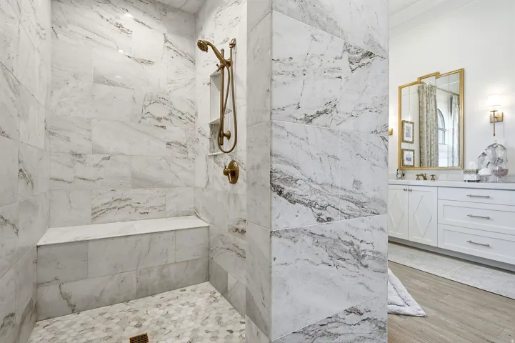 Spacious shower featuring full-height marble-look tile, a built-in bench, and a bronze-finish shower system