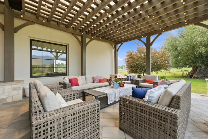 Expansive outdoor patio featuring a wood pergola with string lighting, stone veneer accents, and a large black-framed window