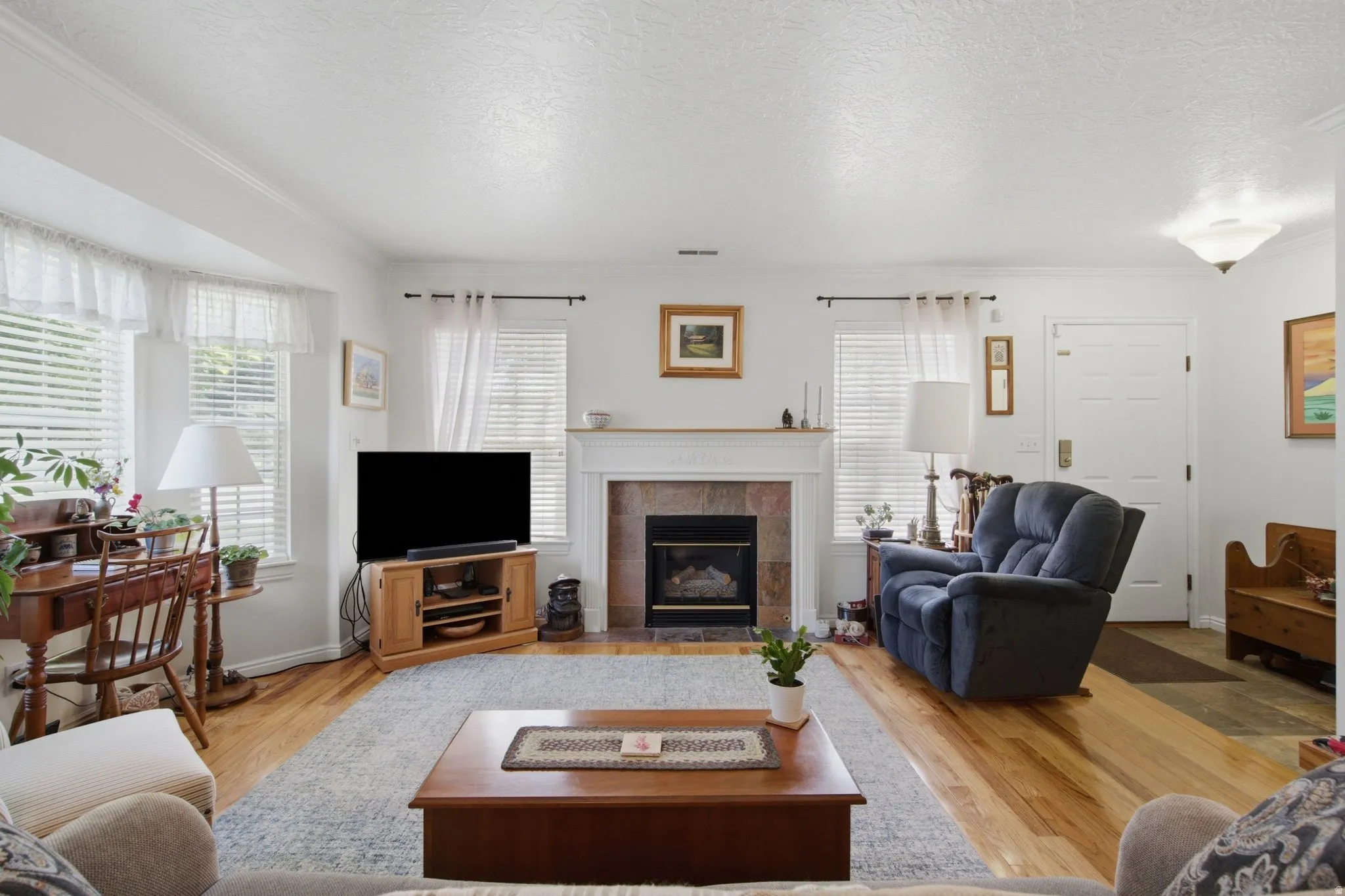 Living room featuring rich wood-finish flooring, a fireplace with a stone surround and a white mantel, and multiple windows with blinds