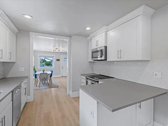 Contemporary kitchen featuring light wood-finish flooring, white shaker-style cabinetry, stainless steel appliances, grey countertops, and a white tile backsplash