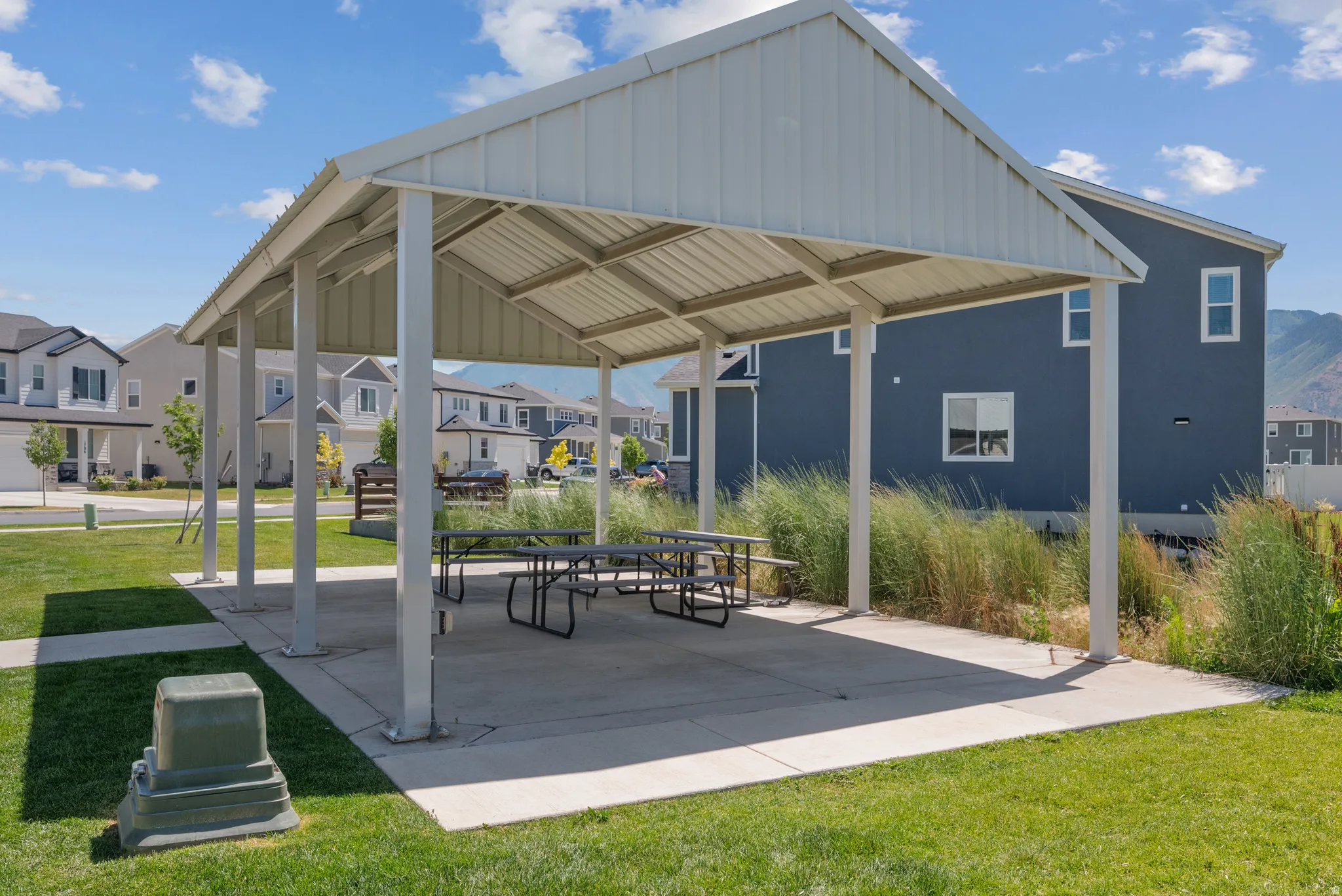 Outdoor covered pavilion with a metal roof and concrete pad, featuring multiple picnic tables and surrounded by green grass