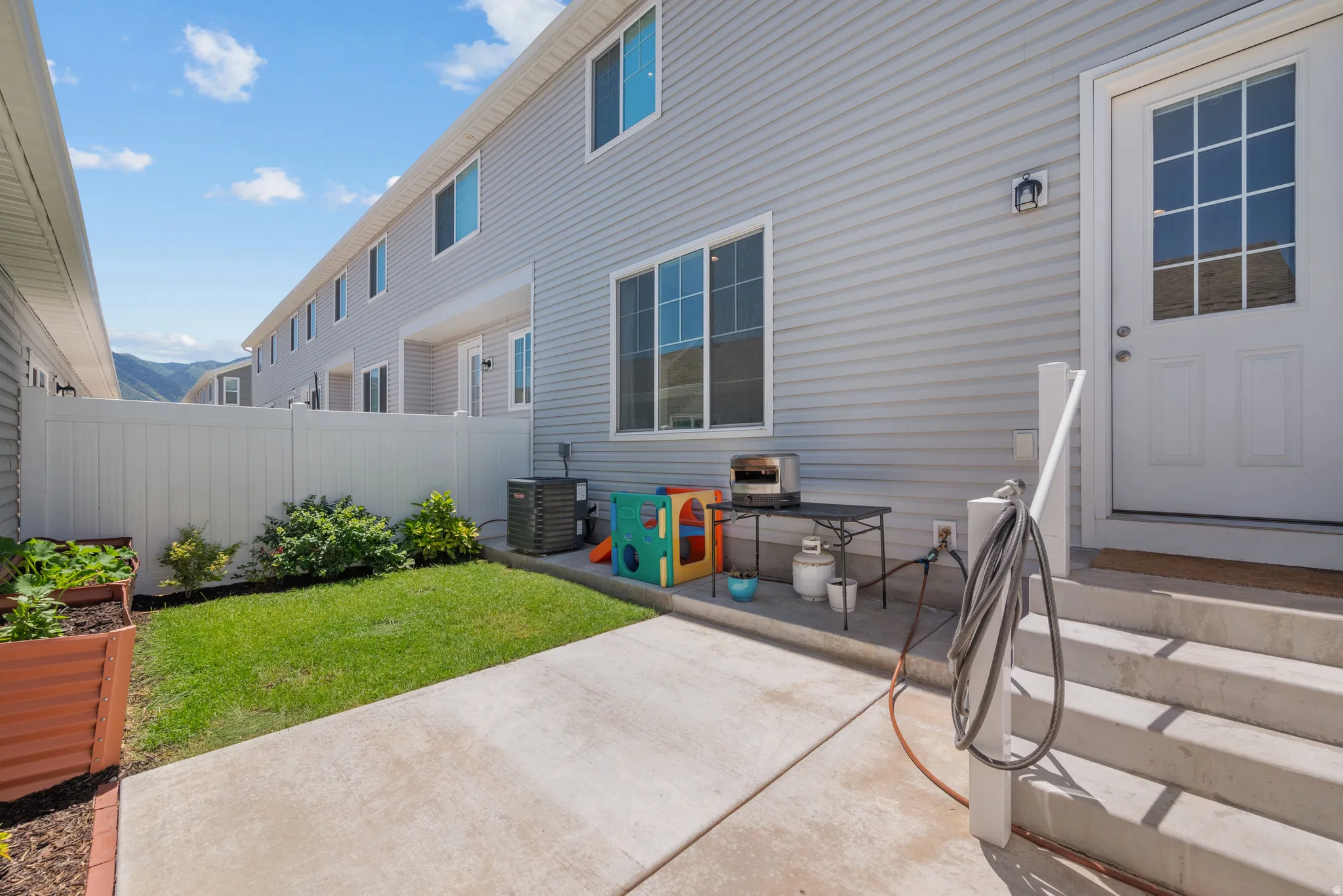 Rear exterior featuring light gray siding, white privacy fencing, concrete patio, grass lawn, and a raised planter box