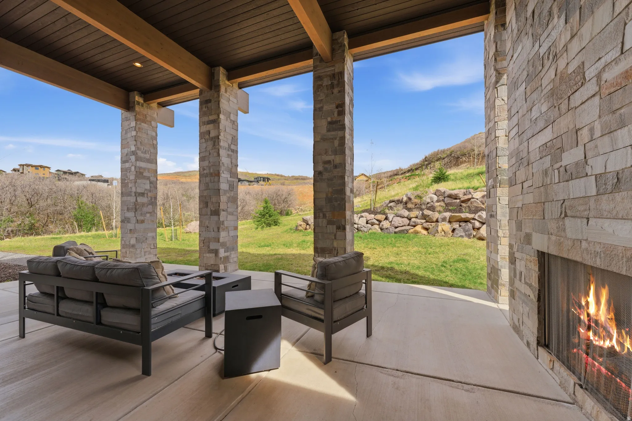 Covered patio featuring a built-in stone fireplace, exposed wood ceiling beams, stone-clad pillars, and a concrete patio surface