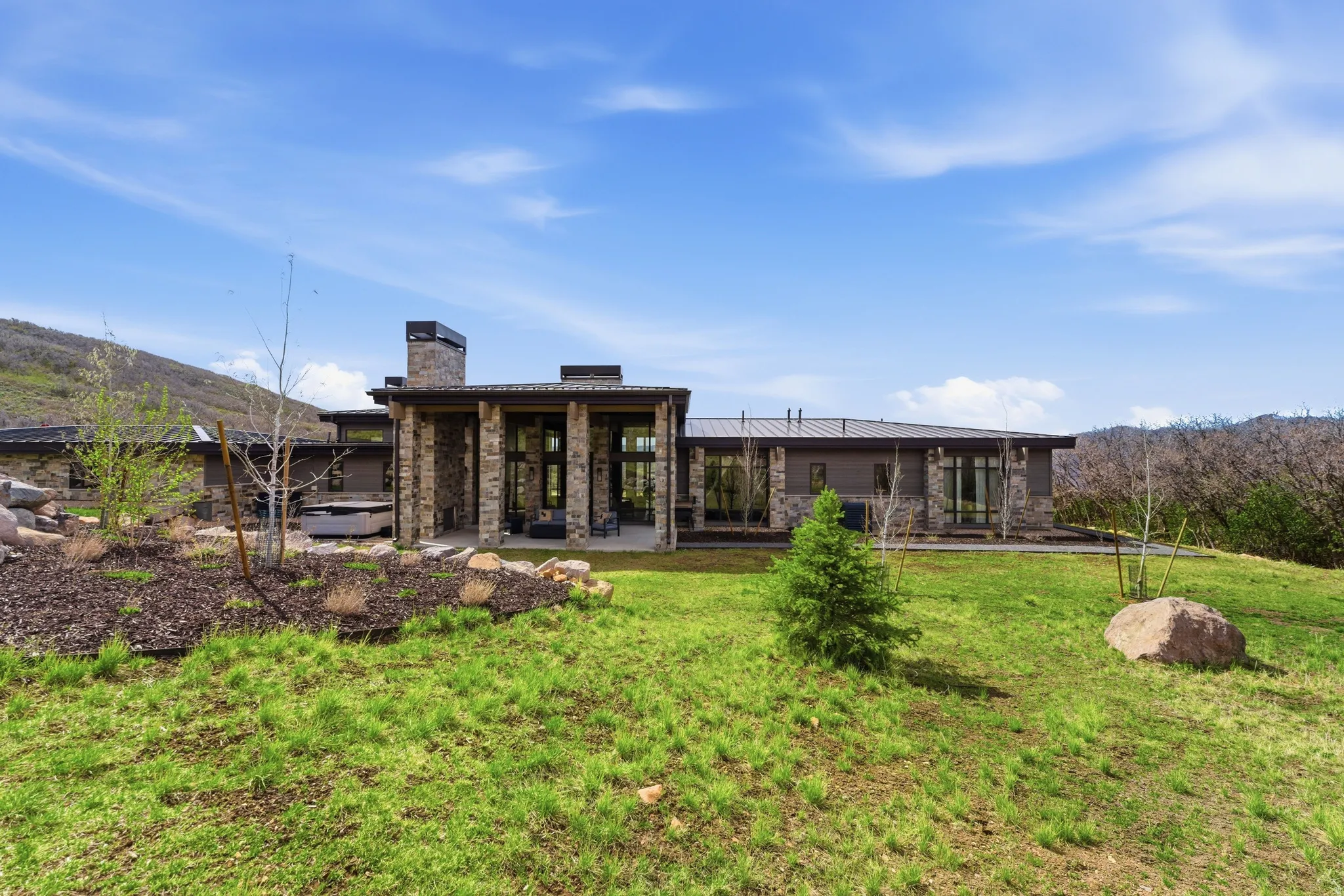 Contemporary architecture featuring a stone and stucco exterior, prominent stone columns, expansive windows, a covered patio, and a stone chimney