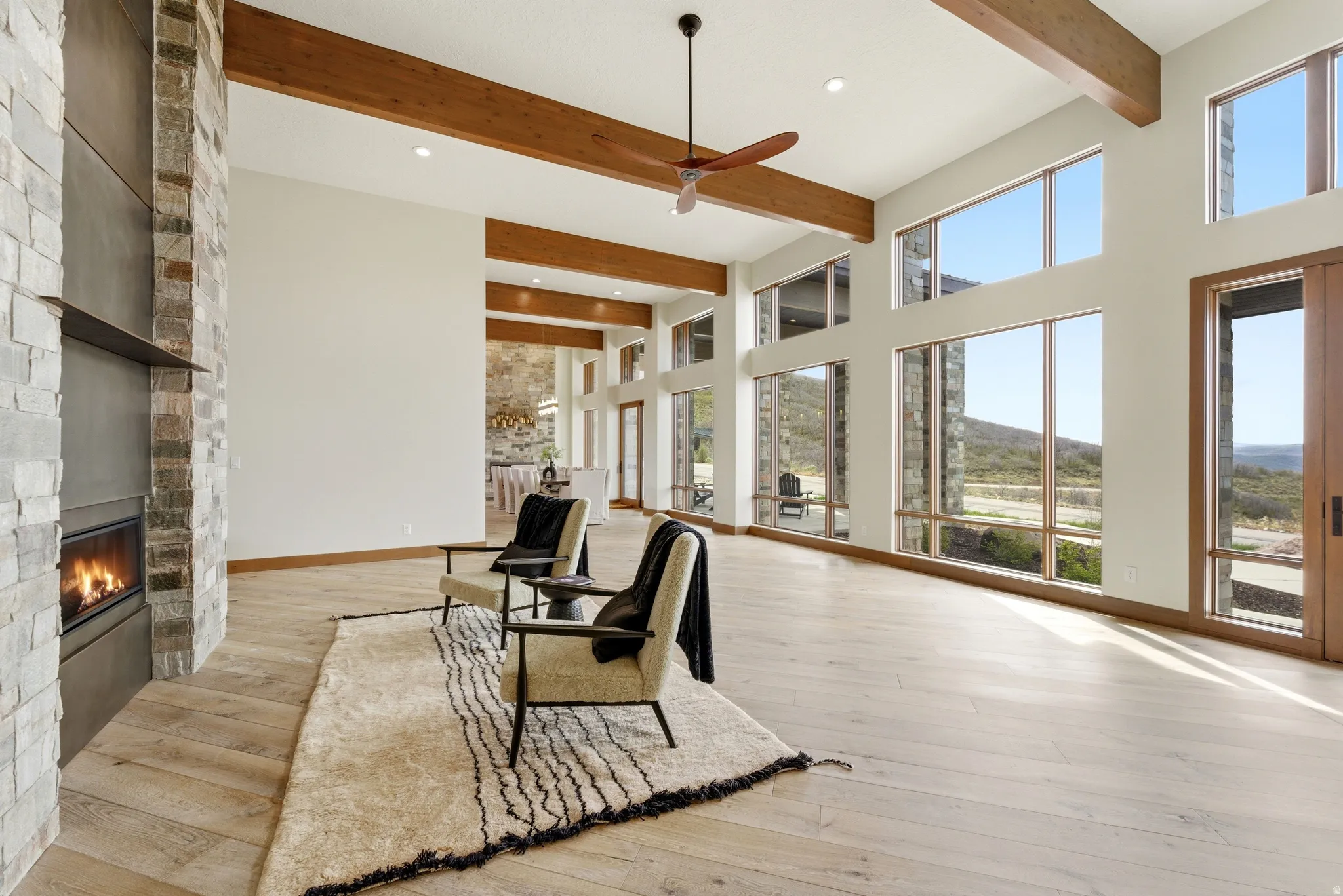 Open-concept living area featuring high ceilings with exposed wood beams, expansive windows, a contemporary fireplace with a stone and dark metal surround, and light wood-finish flooring