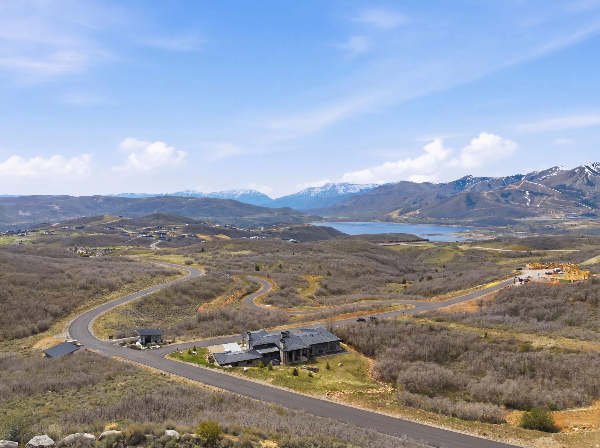 Extensive undeveloped grounds with a winding paved road, a contemporary single-story residence with a dark roof, and distant snow-capped mountains framing a body of water