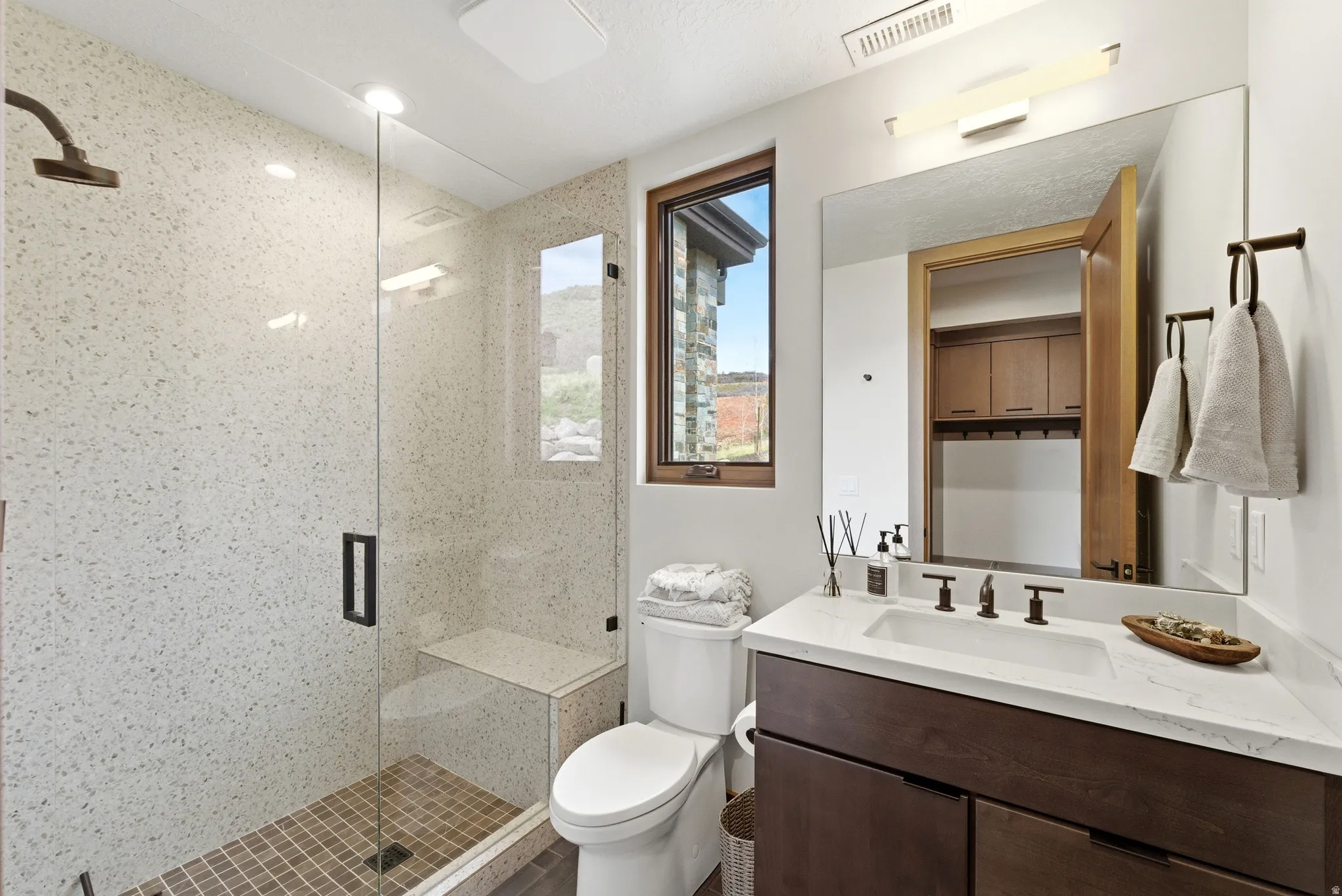 Bathroom featuring a glass-enclosed shower with a built-in bench, a wood-framed window, a dark wood vanity with a white countertop and integrated sink, and an expansive wall mirror