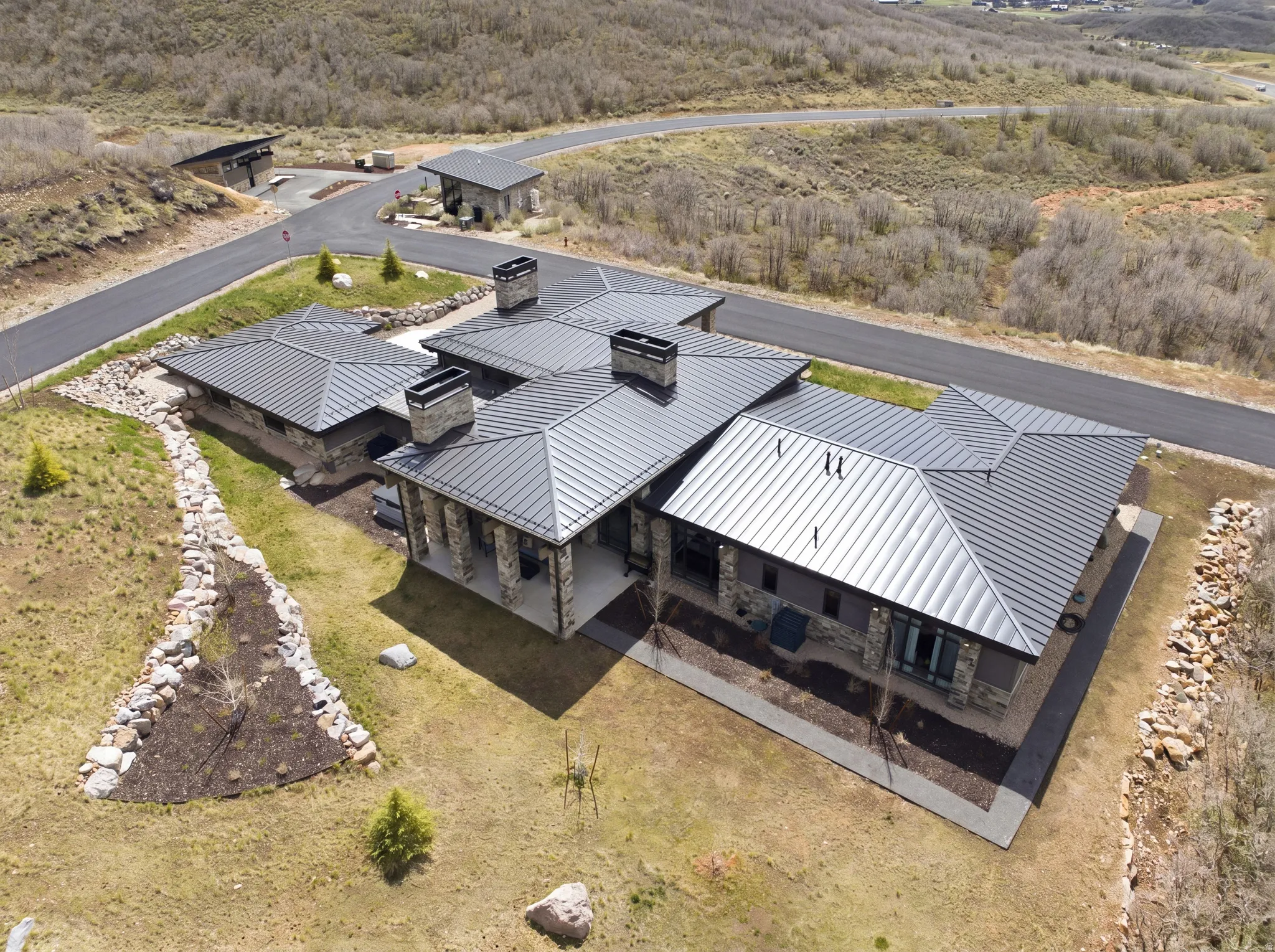 Expansive modern residence featuring a dark metal standing seam roof, stone and stucco exterior, multiple prominent chimneys, and covered outdoor living spaces