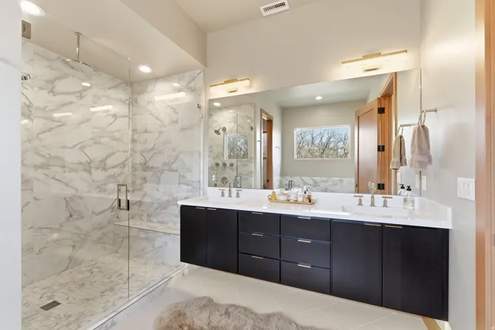 Spacious bathroom featuring a frameless glass shower enclosure with marble-finish tile, a built-in shower bench, and a dual vanity with dark wood-finish cabinetry, white countertops, and undermount sinks