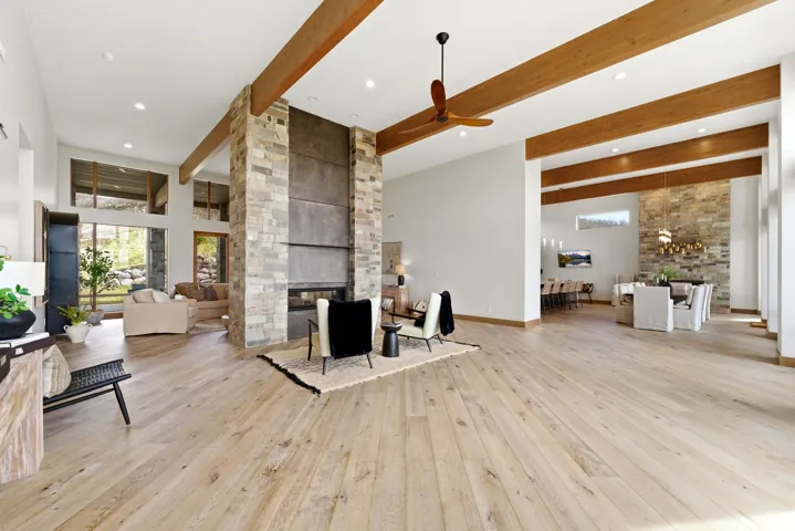 Expansive living area featuring light wood-finish flooring, a stone-clad fireplace with a dark metal surround, exposed wood ceiling beams, and recessed lighting