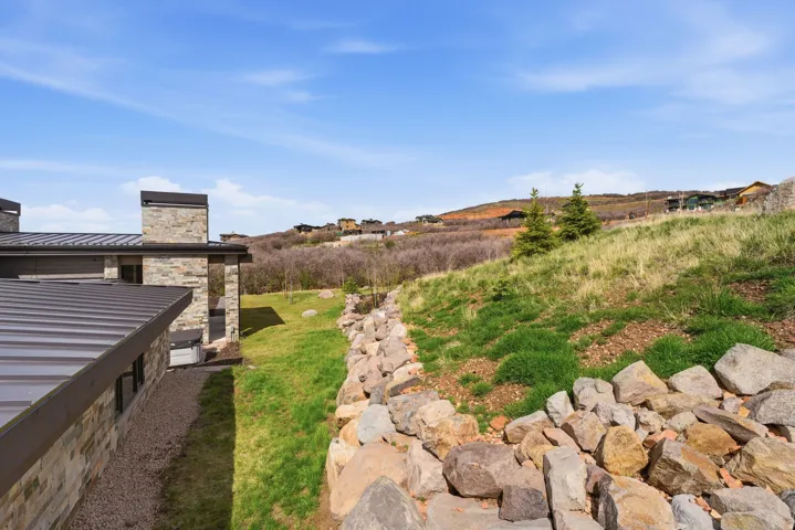 Stone and corrugated metal exterior with a prominent stone chimney, surrounded by a rock retaining wall and sloped grassy terrain