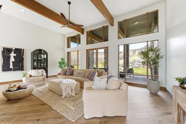Spacious living area featuring high ceilings with exposed wood beams, extensive floor-to-ceiling windows, and wood-finish flooring