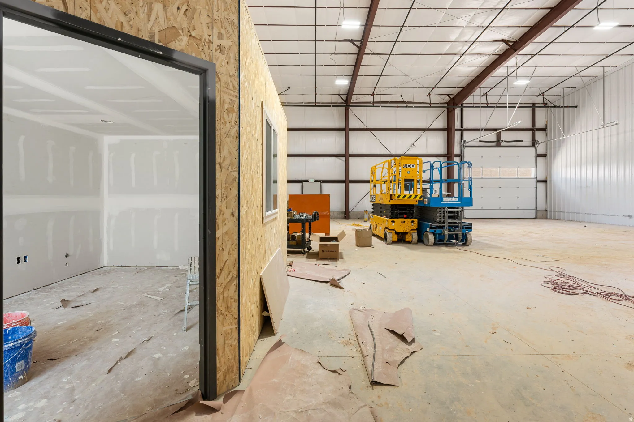 Spacious commercial interior featuring a large roll-up door, exposed steel beam construction, concrete flooring, and an unfinished office space with drywall-ready walls