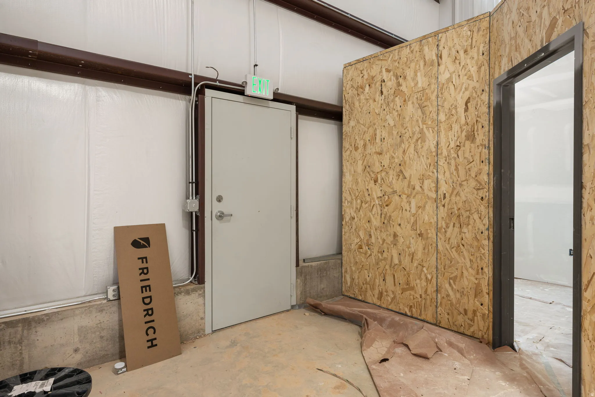 Interior space with a concrete floor, light-toned insulated panel wall, exposed brown structural beam, and an interior door with a grey finish