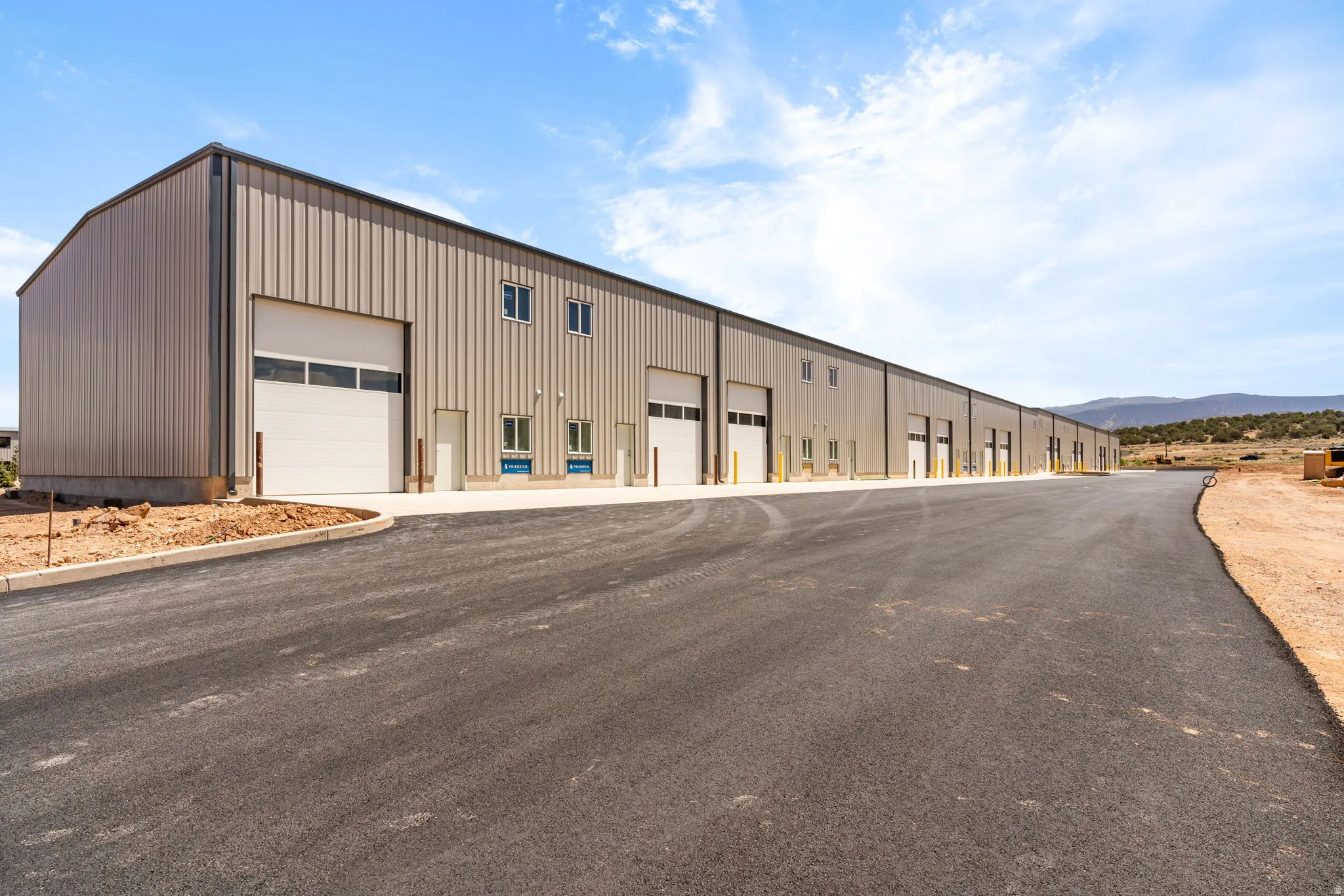 Expansive light tan corrugated metal industrial building featuring multiple roll-up garage doors, ground-level entry doors, and upper-level windows