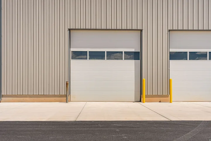 Industrial building featuring vertical metal siding, two overhead doors with window panels, a concrete approach, and asphalt paving