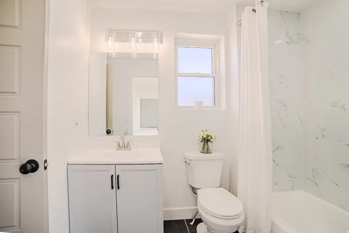 Bathroom featuring a white vanity with a single sink, an overhead vanity light, a toilet, and a shower-tub combination with white marble-look tile surround