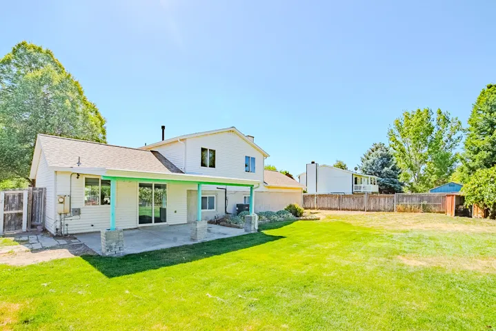 Expansive backyard featuring a large concrete patio, covered patio area with support columns, white siding exterior, and mature perimeter trees