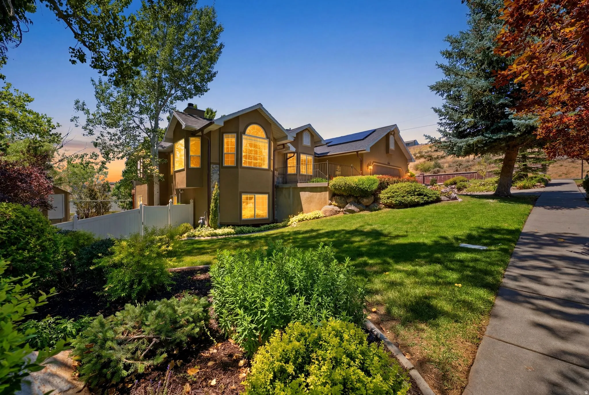 Multi-story residence featuring a landscaped front yard with established shrubs and trees, a concrete pathway, a white privacy fence, and a visible garage