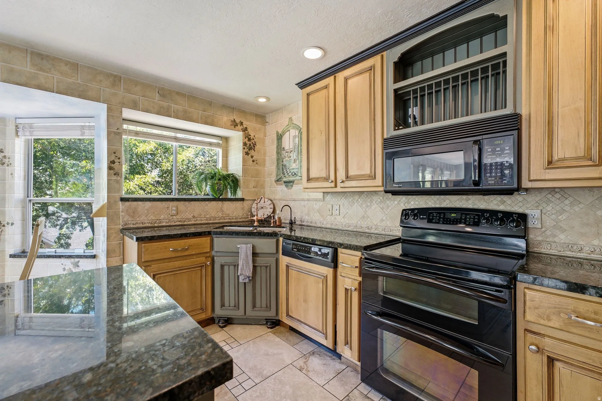 Kitchen featuring stone-look tile flooring, light wood-finish cabinetry, black granite countertops, and a black appliance suite