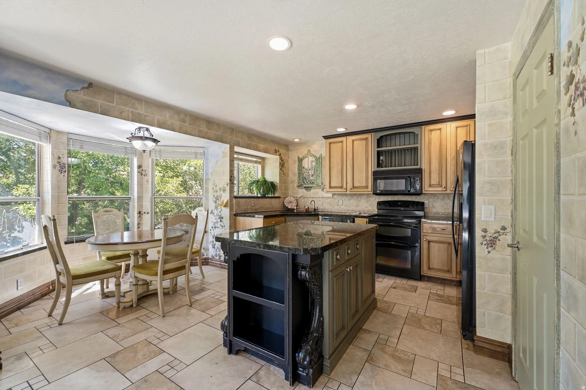 Kitchen featuring a multi-toned tile floor, a central island with a dark countertop and carved details, wood-finish cabinetry, built-in appliances, and a bay window dining area with a flush-mount light fixture