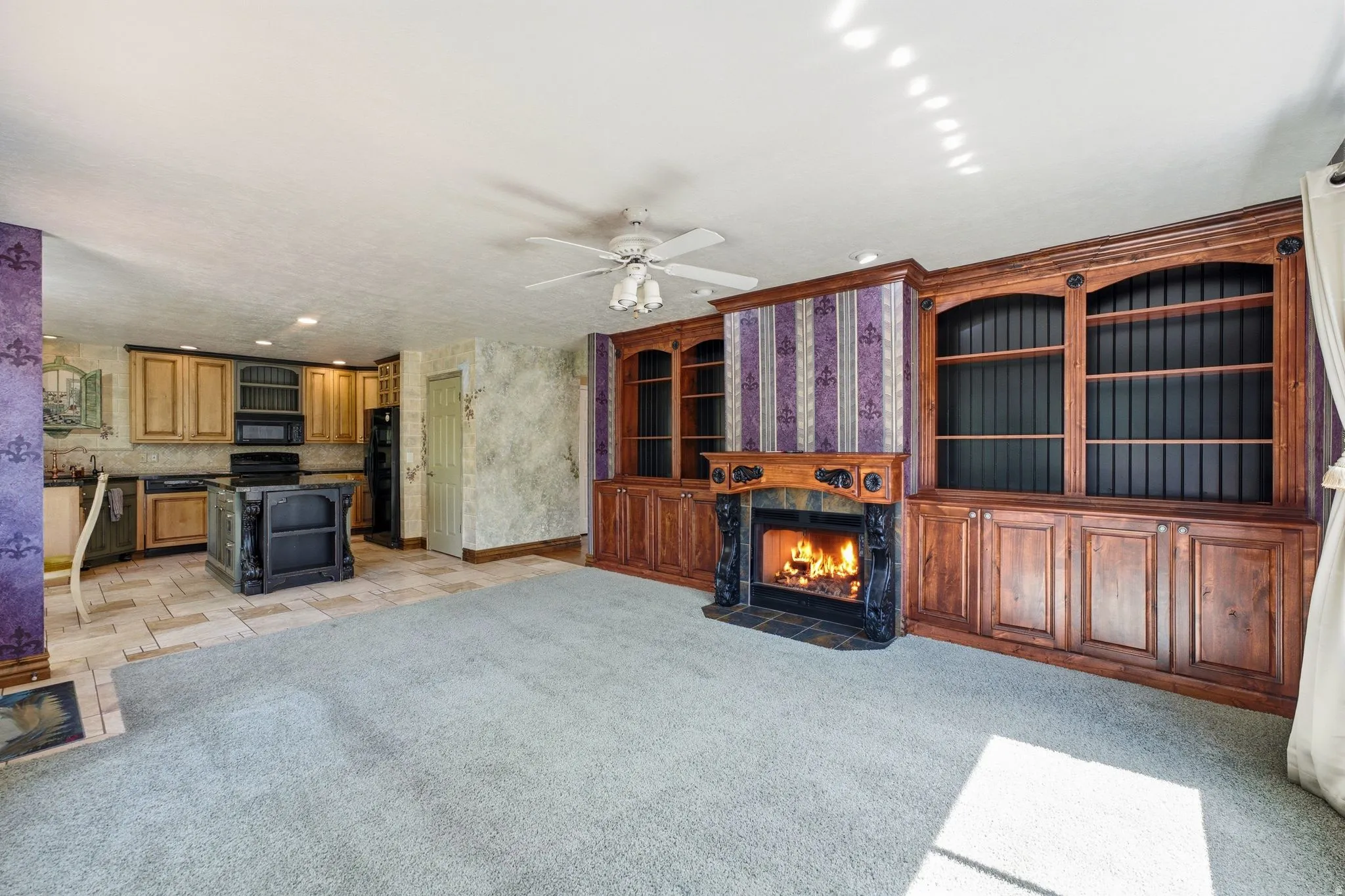 Built-in wood cabinetry with shelving and base storage, integrated fireplace with a stone hearth and carved mantel, kitchen featuring wood cabinetry and an island, tile flooring in the kitchen area, and a ceiling fan with integrated lighting