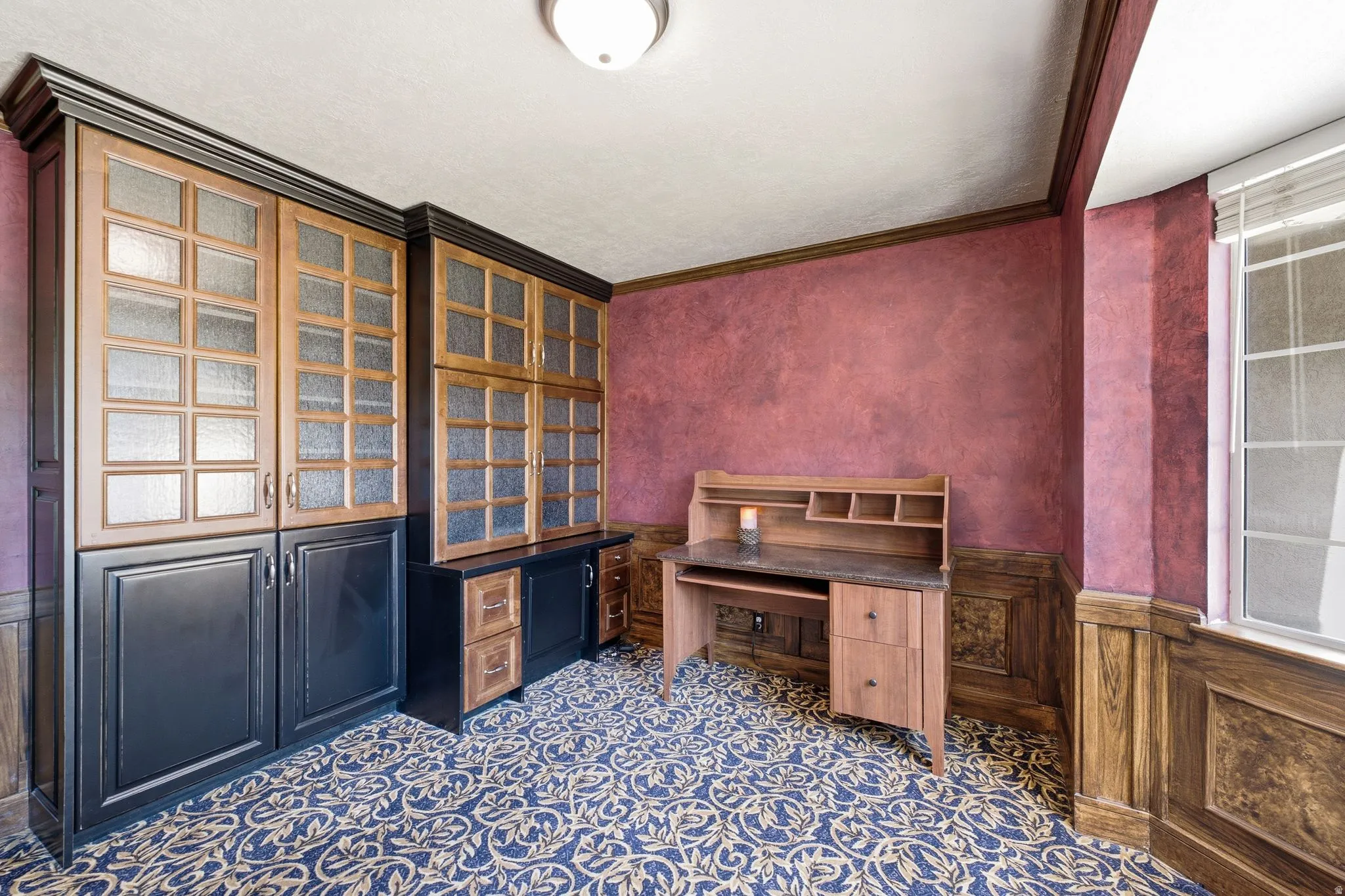 Custom built-in cabinetry with glass-front uppers, a built-in desk with a stone-finish countertop, decorative wainscoting, and patterned carpet