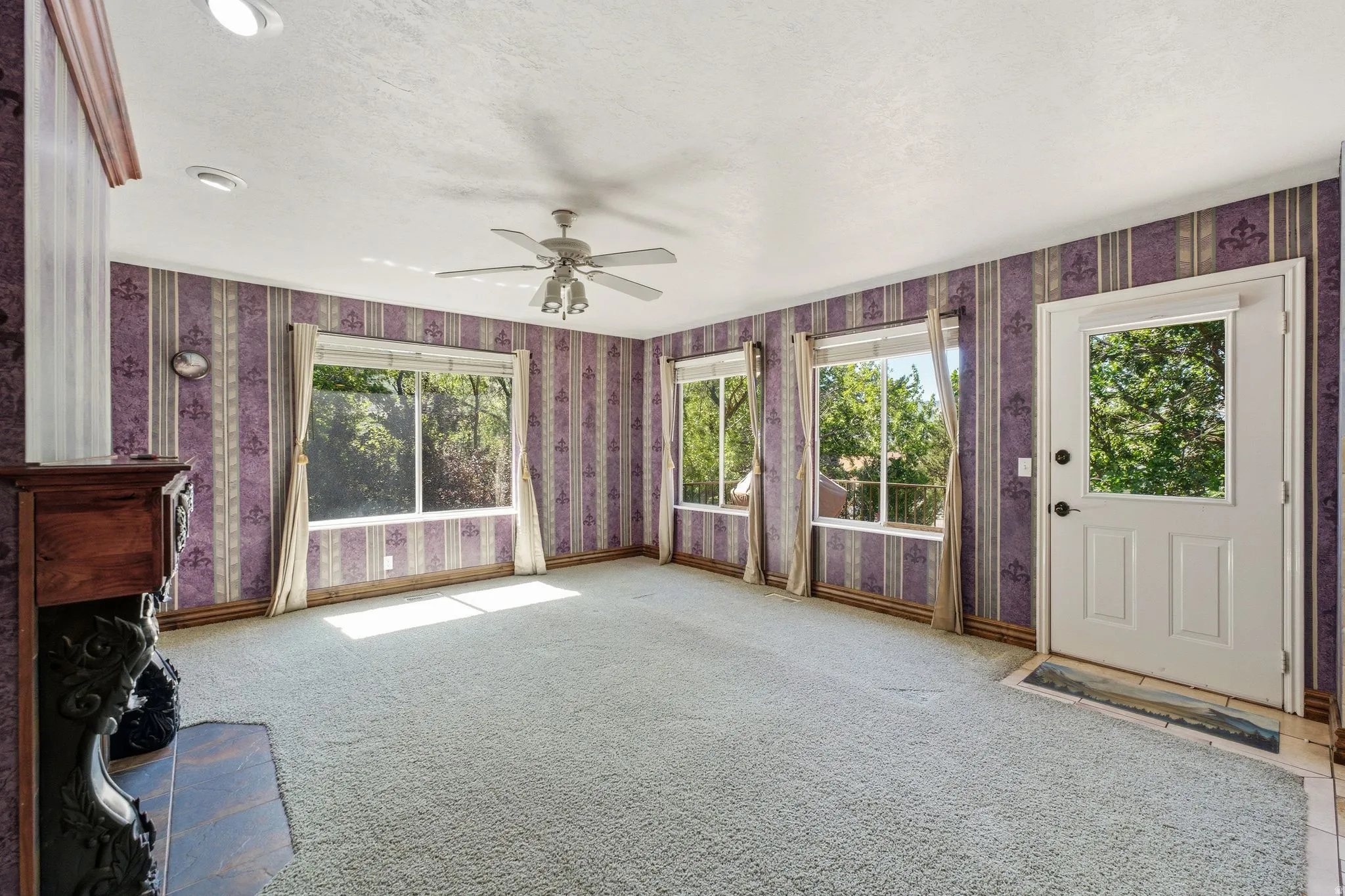 Living area featuring multiple windows, a ceiling fan, patterned wallpaper, and a fireplace with a dark wood mantel