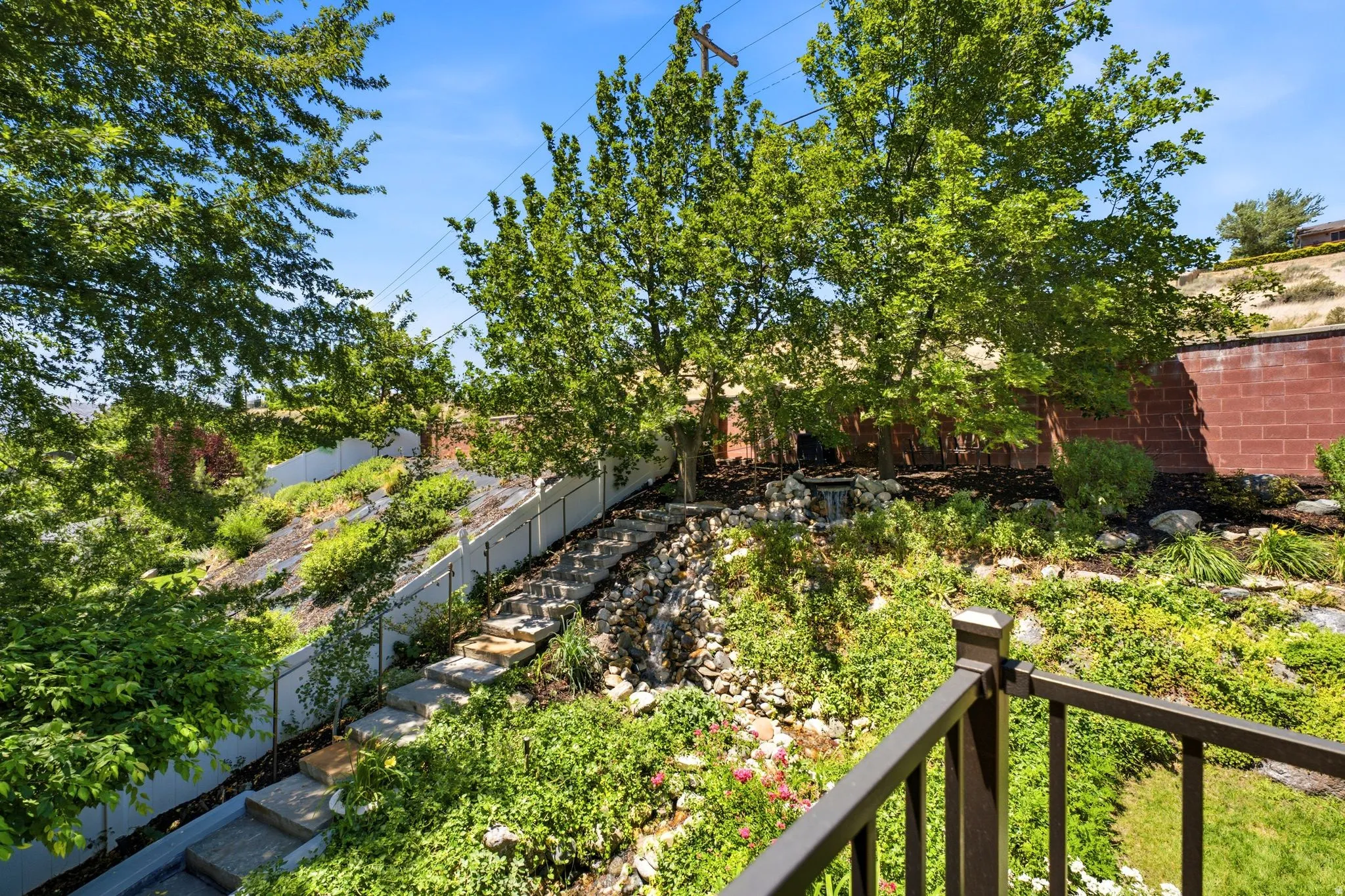 Tiered landscaping featuring stone steps, mature trees, a white perimeter fence, and a rock water feature