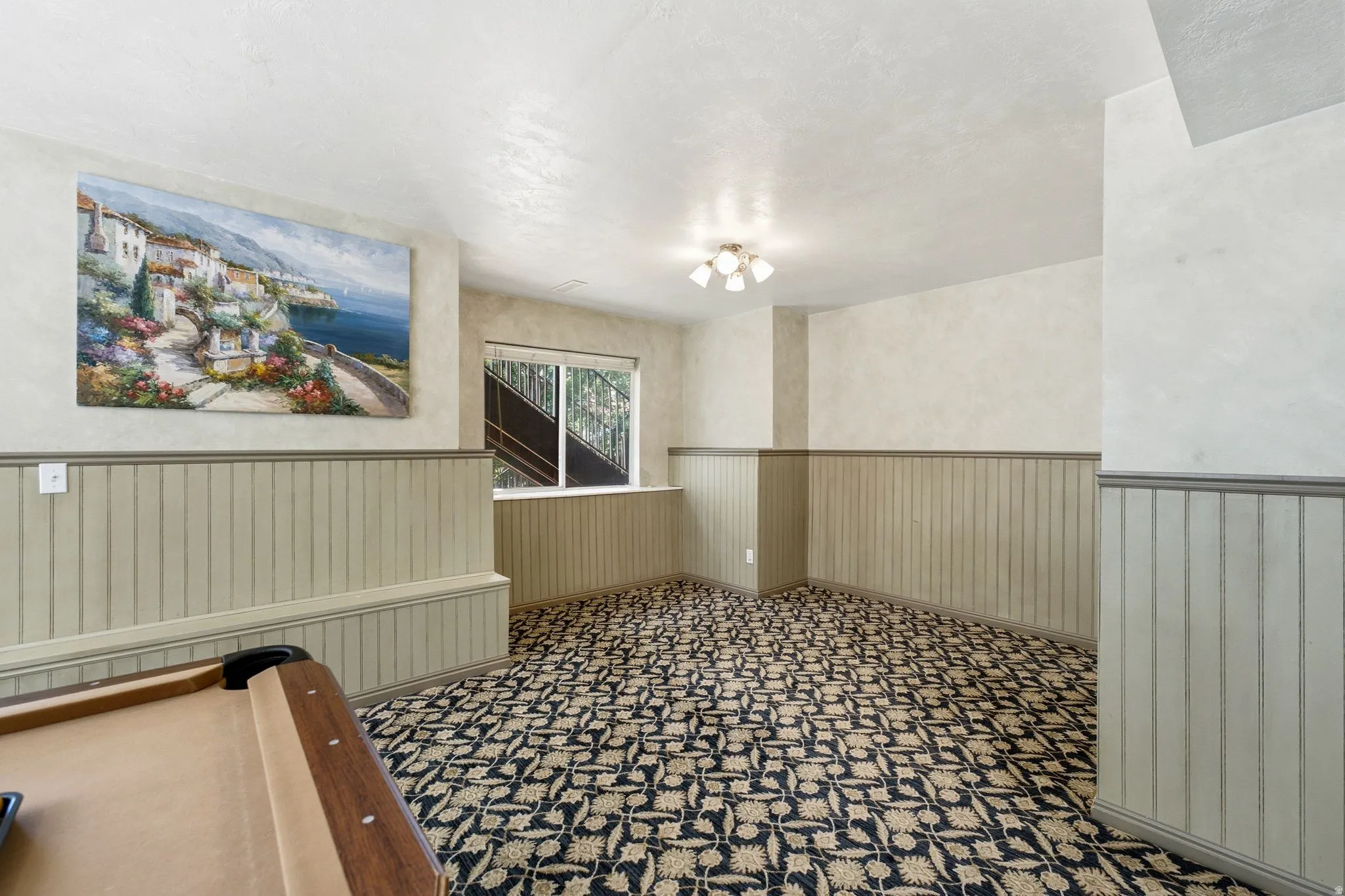 Recreational space featuring patterned carpet flooring, beadboard wainscoting, and a window with a dark metal railing visible