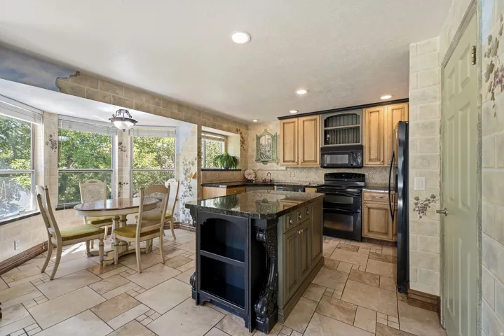 Kitchen featuring a multi-toned tile floor, a central island with a dark countertop and carved details, wood-finish cabinetry, built-in appliances, and a bay window dining area with a flush-mount light fixture