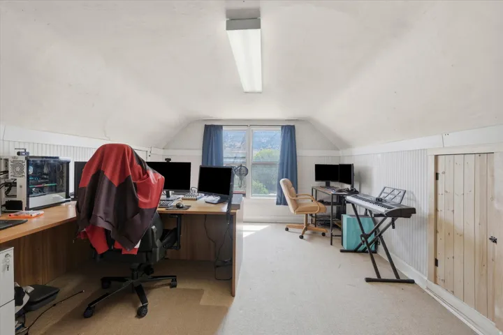 Attic space featuring sloped ceilings, a prominent window with blue drapes, and a built-in storage door with vertical wood paneling