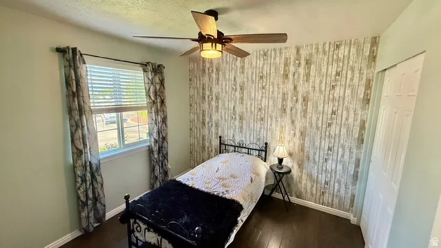 Wood-finish flooring, accent wall with vertical paneling, single window with blinds, ceiling fan with integrated lighting, and a bi-fold door