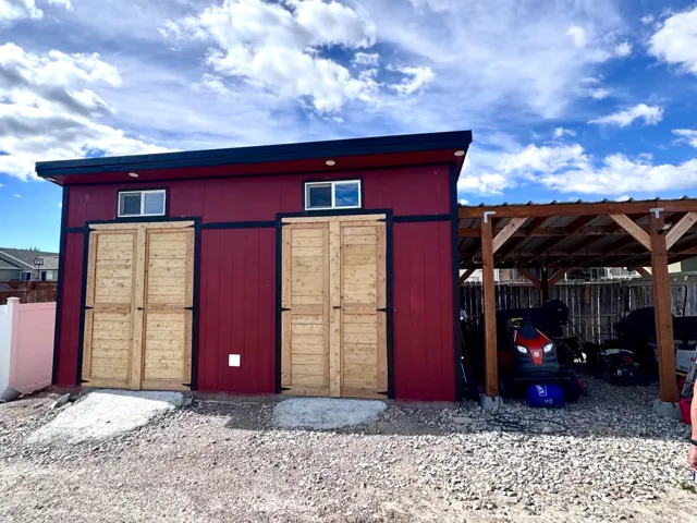 Red-sided outbuilding with two wooden barn-style doors, individual concrete pads, and upper transom windows