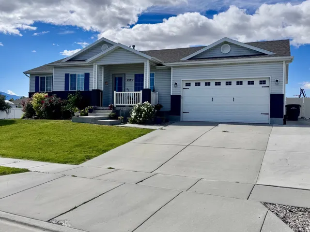 Ranch-style residence featuring light-colored siding with dark trim accents, a prominent front porch with white railings, a two-car garage with paneling and windows, a well-maintained lawn, and a paved driveway