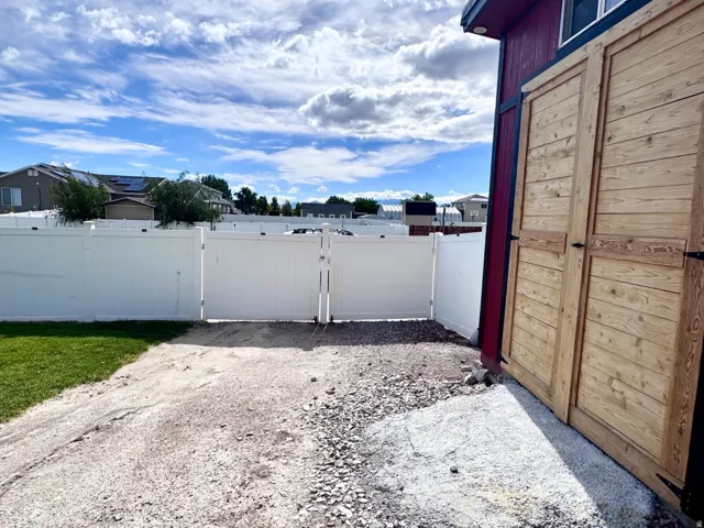 Gravel driveway with a white vinyl fence, double gate, and a red structure featuring wooden doors and a window