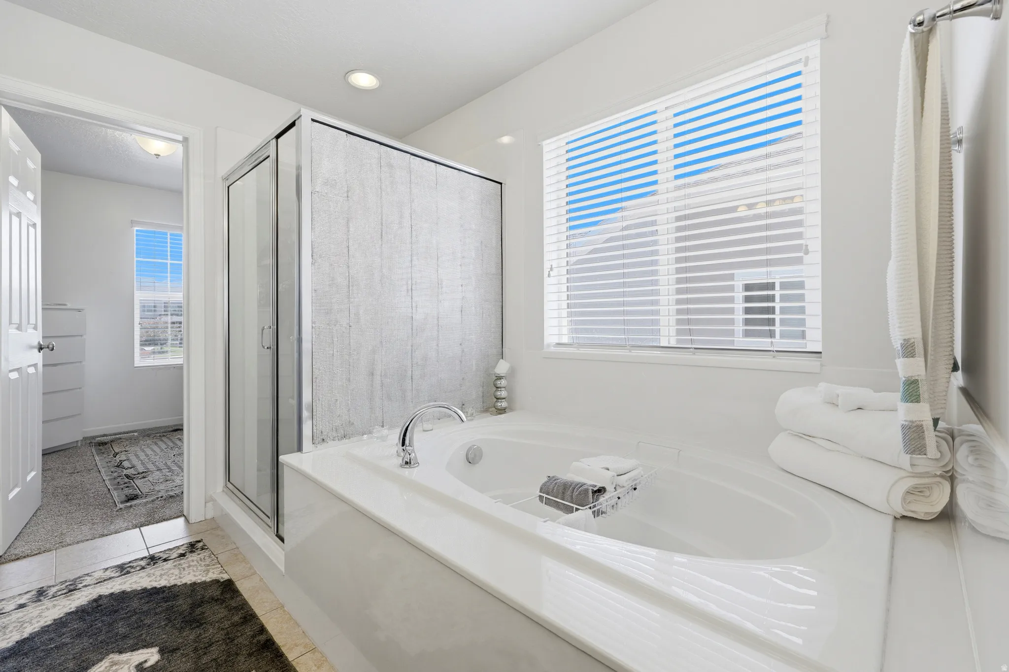 Bright bathroom featuring a built-in soaking tub, separate glass-enclosed shower with textured wall, tile flooring, and a large window with blinds