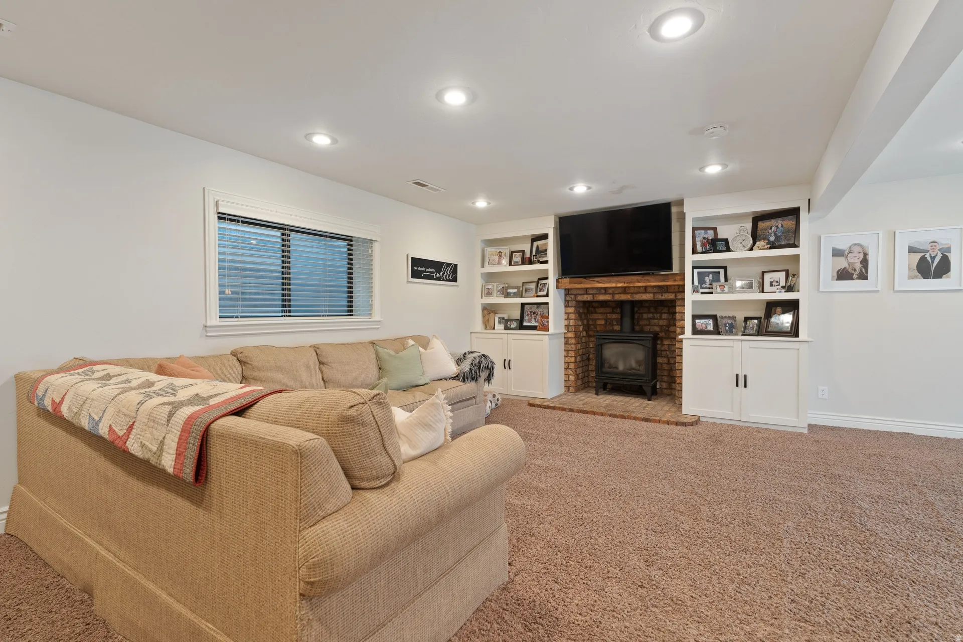 Living area featuring a brick-surround fireplace with a black wood-burning stove, built-in shelving, white cabinetry, recessed lighting, and a light-colored window with blinds