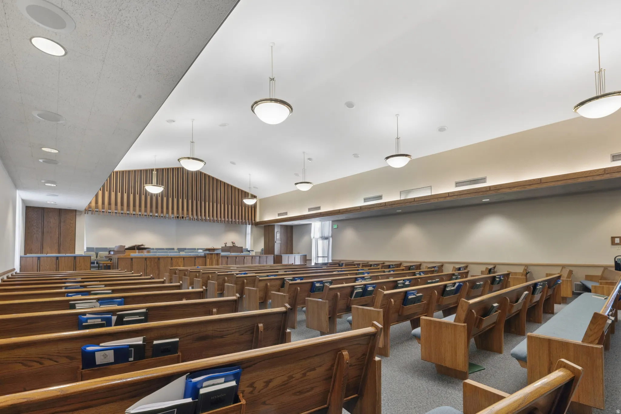 Spacious interior featuring rows of wood benches, acoustic paneling, suspended globe pendant lighting, recessed ceiling fixtures, and neutral-toned walls