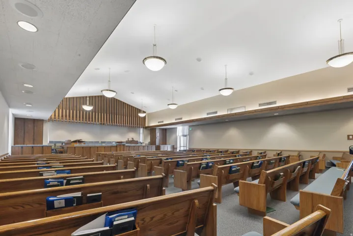 Spacious interior featuring rows of wood benches, acoustic paneling, suspended globe pendant lighting, recessed ceiling fixtures, and neutral-toned walls