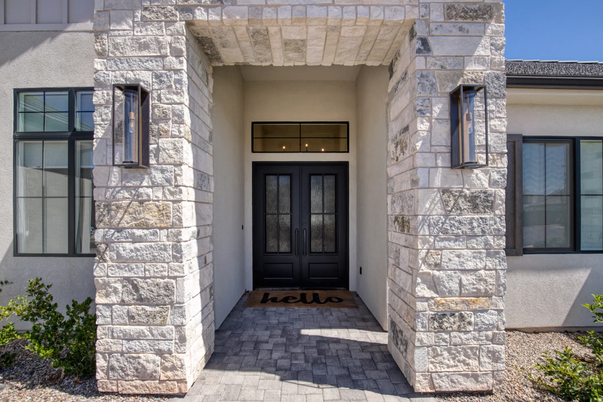 Stone-clad entryway featuring a double-door entrance with decorative glass inserts, an overhead transom window, and modern exterior sconces