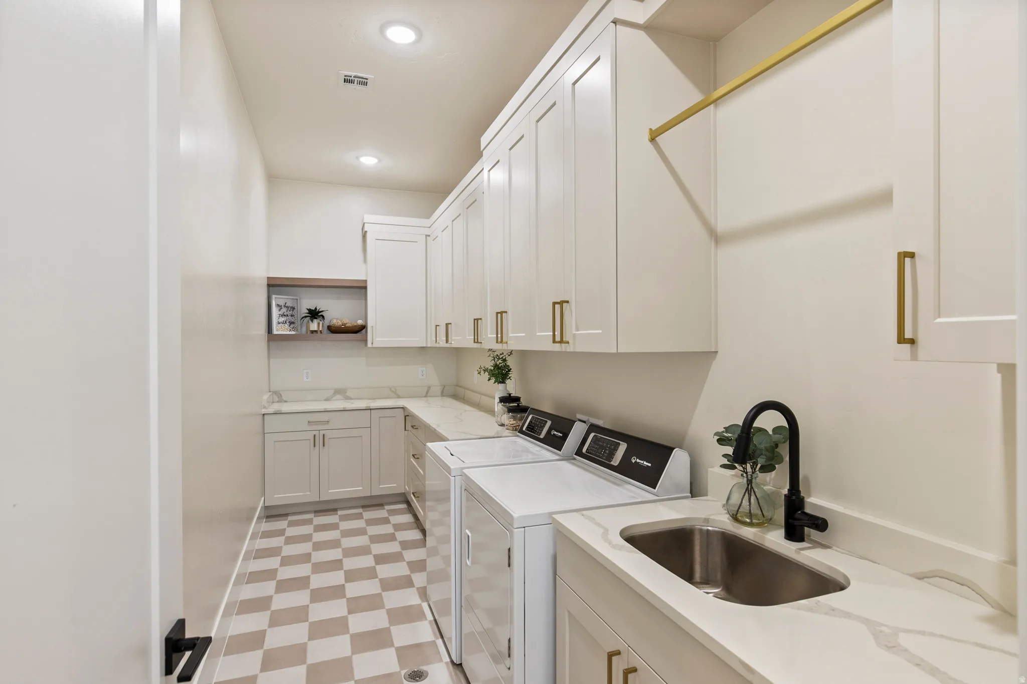 Laundry room featuring checkerboard tile flooring, extensive white cabinetry with gold hardware, light-toned countertops with veining, a stainless steel sink with a matte black faucet, and an overhead drying rack