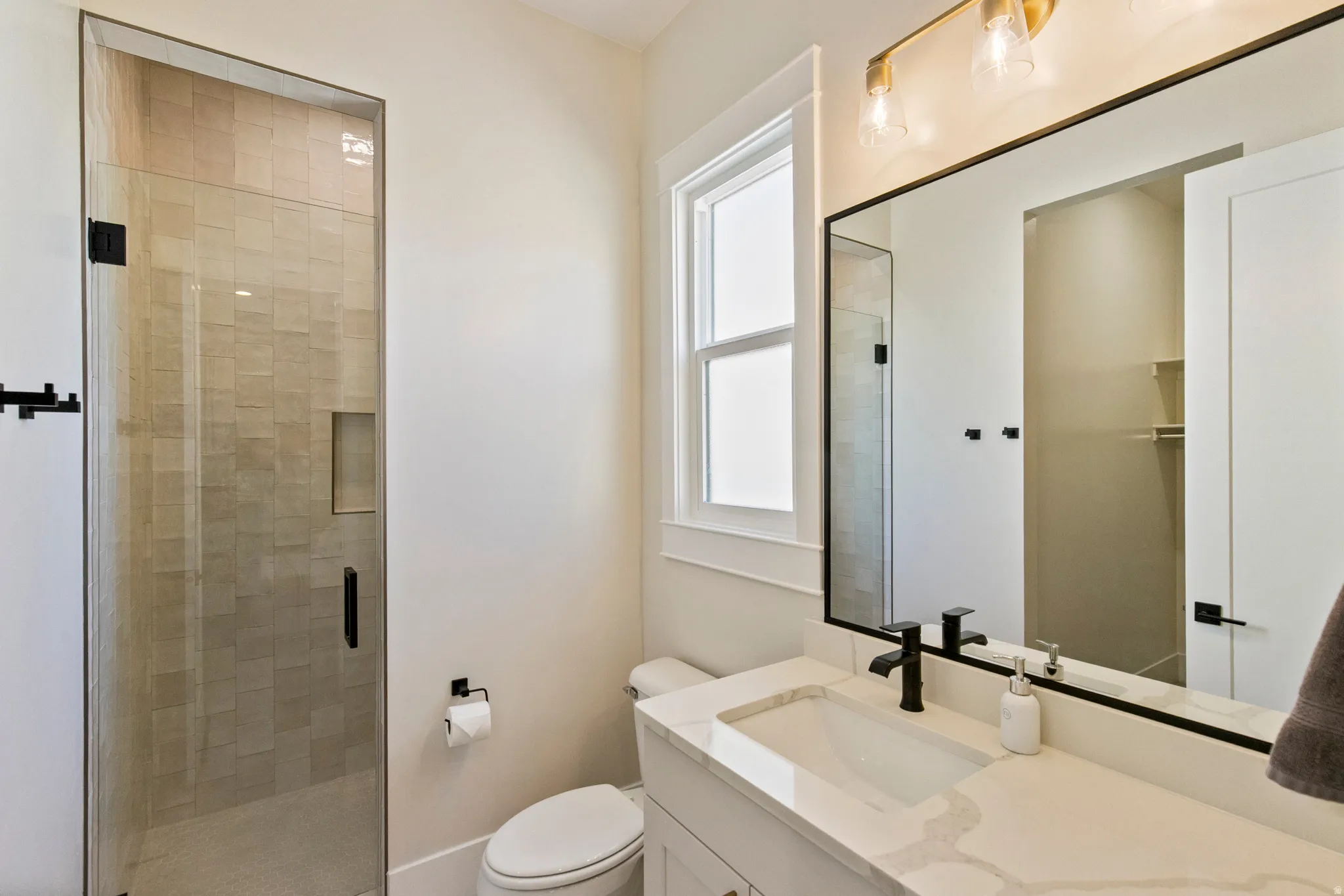 Bathroom featuring a frameless glass shower enclosure with tiled walls and a recessed niche, a white vanity with an integrated rectangular sink and stone countertop, a large mirror with a black frame, and matte black fixtures