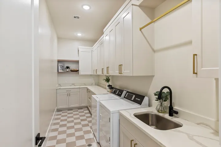 Laundry room featuring checkerboard tile flooring, extensive white cabinetry with gold hardware, light-toned countertops with veining, a stainless steel sink with a matte black faucet, and an overhead drying rack