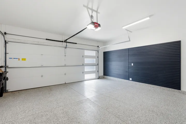 Attached two-car garage featuring a light gray epoxy floor coating, white overhead garage door with frosted glass panels, and a dark slatwall storage system