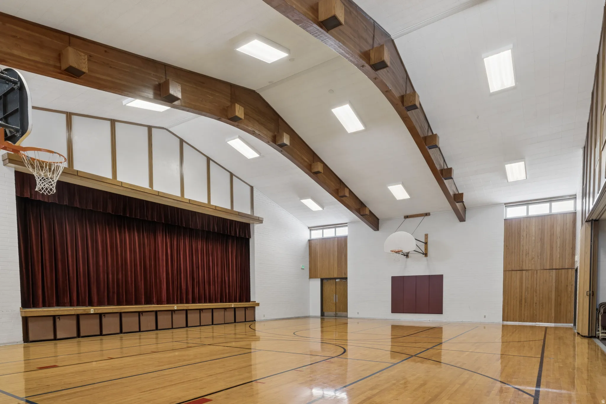 Gymnasium featuring a polished hardwood court, exposed wood beams, and a vaulted ceiling with integrated lighting. Stage with curtain on adjacent wall.
