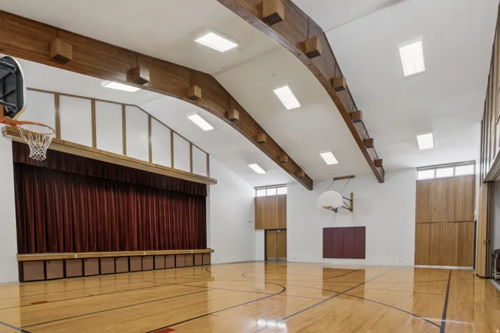 Gymnasium featuring a polished hardwood court, exposed wood beams, and a vaulted ceiling with integrated lighting. Stage with curtain on adjacent wall.